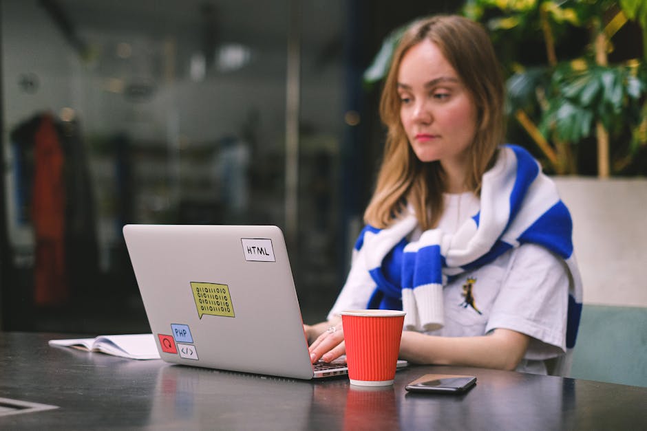 A close-up view of a laptop computer on a wooden desk, displaying lines of colorful code on its screen, with a ceramic coffee mug placed nearby. The mug has a humorous message that reads 'This coffee